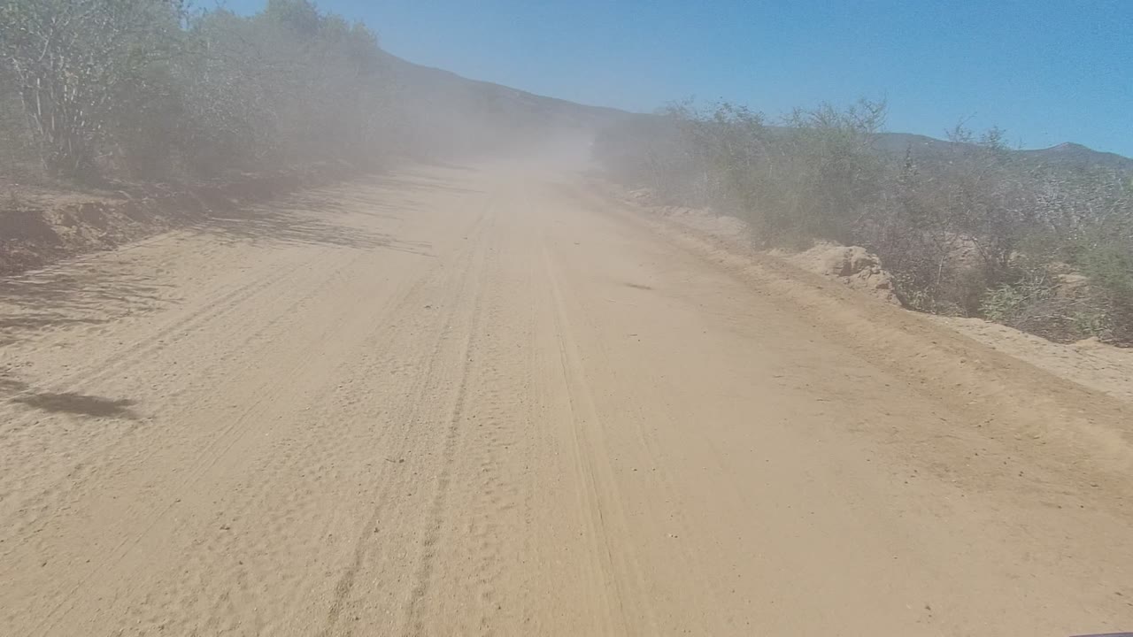At 0:02, the sandy road in Los Cabos bends gently through dry brush as dust softens the mid-distance and the blue sky remains clear, with no visible people.