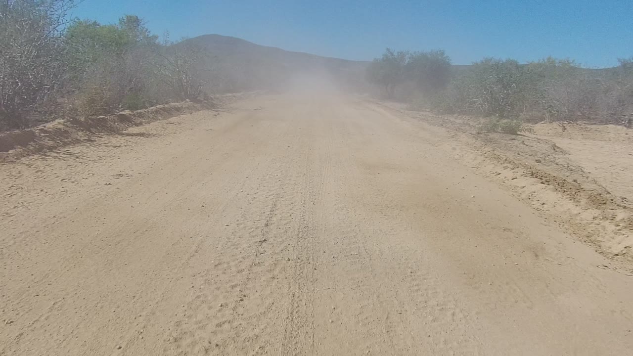 At 0:00, a sandy unpaved road in Los Cabos runs toward a faint dust cloud, with dry brush on both sides, low hills in the distance, and no visible people.
