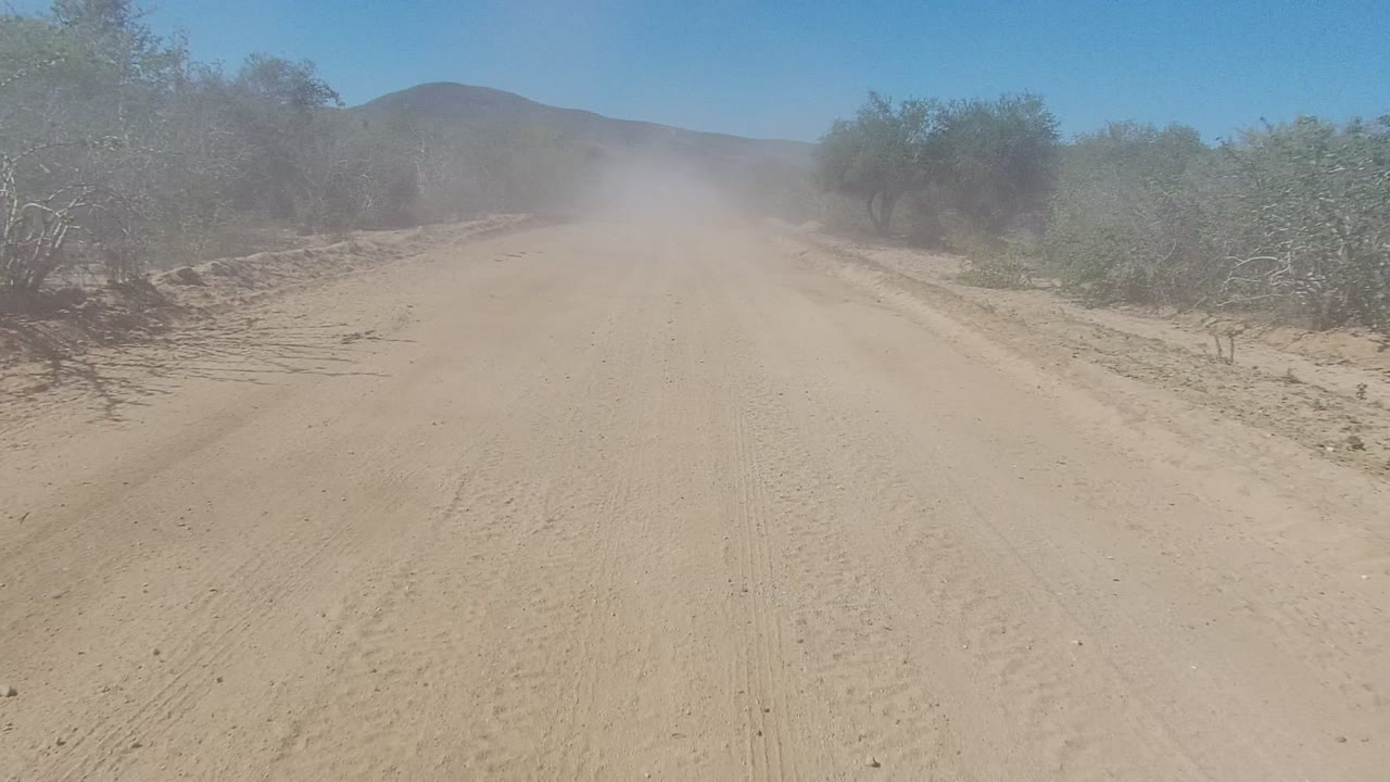 Unpaved desert road with dust drifting ahead and brush along the shoulder.
