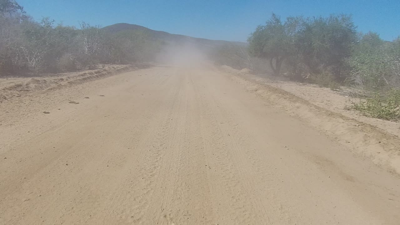 At 0:02, a broad sandy road in Los Cabos leads into a central dust cloud, with dry trees and brush lining the sides and no visible people.