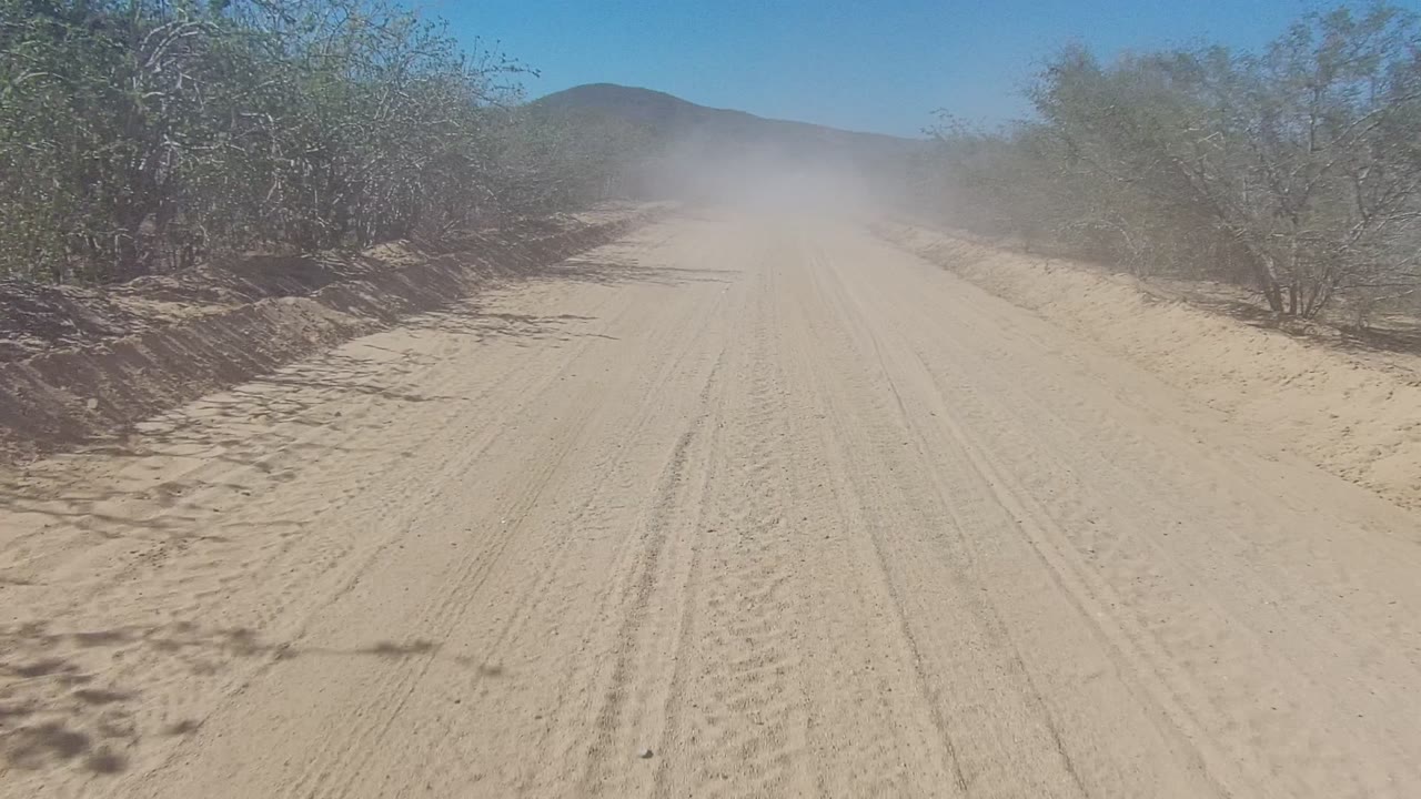 Centered sandy road with tire tracks, roadside brush, and hazy dust in the distance.