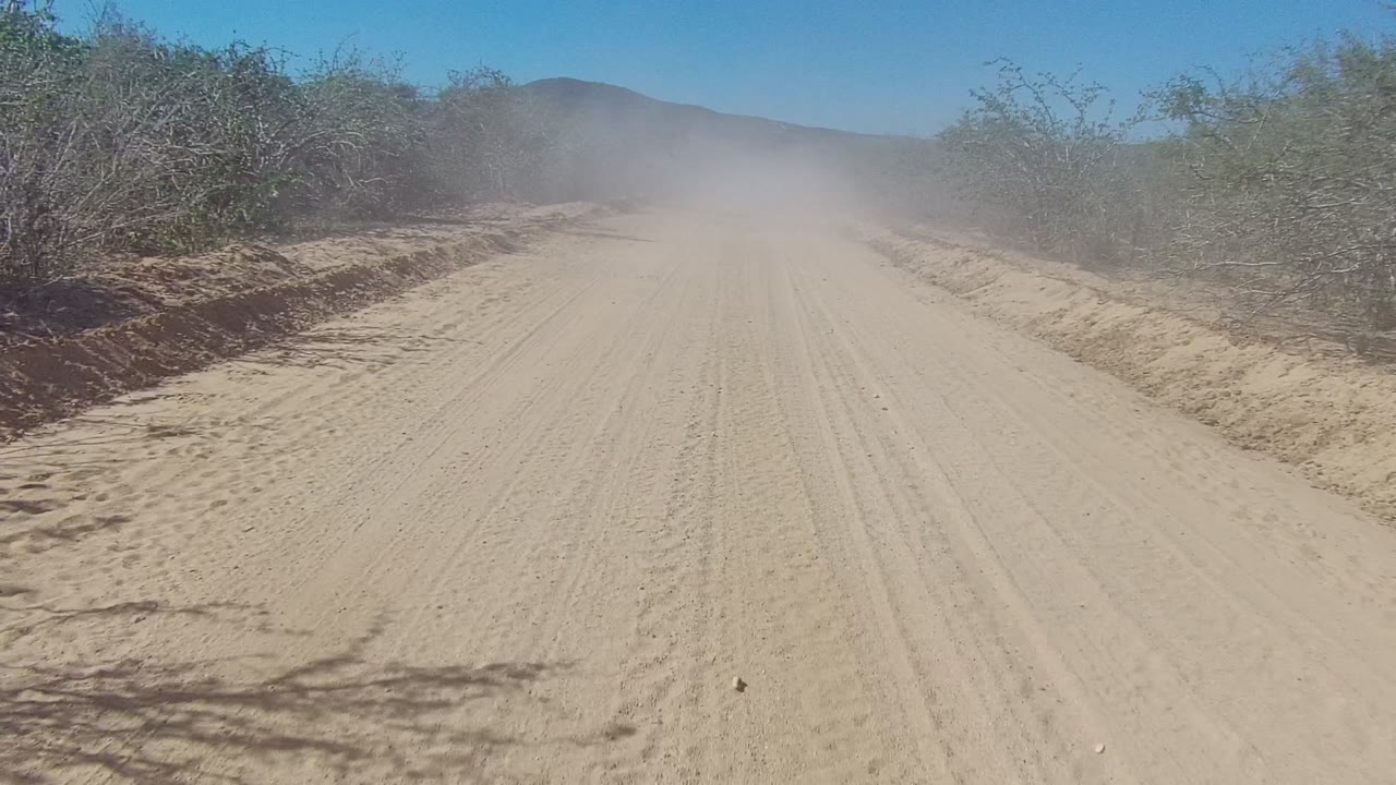 A wider stretch of sandy road in Los Cabos, Baja California Sur leads into a lingering dust cloud, with scrub vegetation, low hills, blue sky, and no visible people.