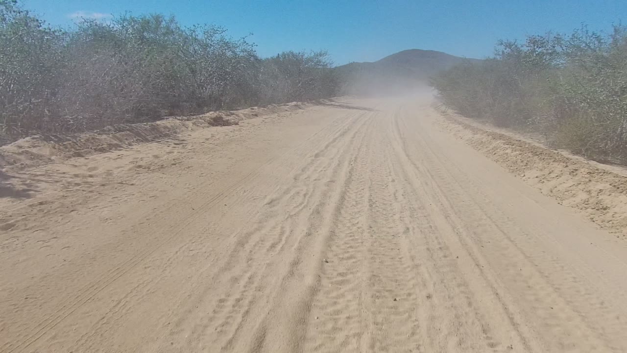 Sandy dirt road in Los Cabos, Mexico with parallel tire tracks, brush on both sides, distant hills, a dust cloud ahead, and no visible people.