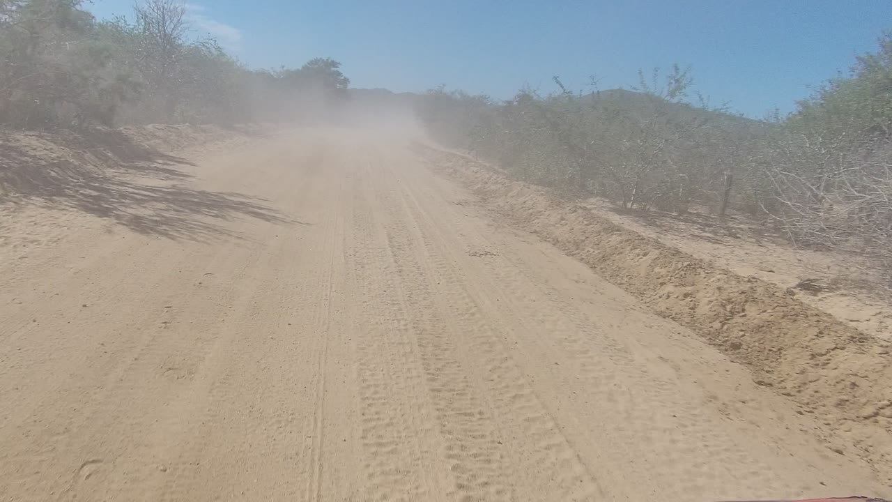 Forward view down a dusty sandy road in Los Cabos, Baja California Sur, with tire tracks, brush on both sides, haze ahead, and no visible people.