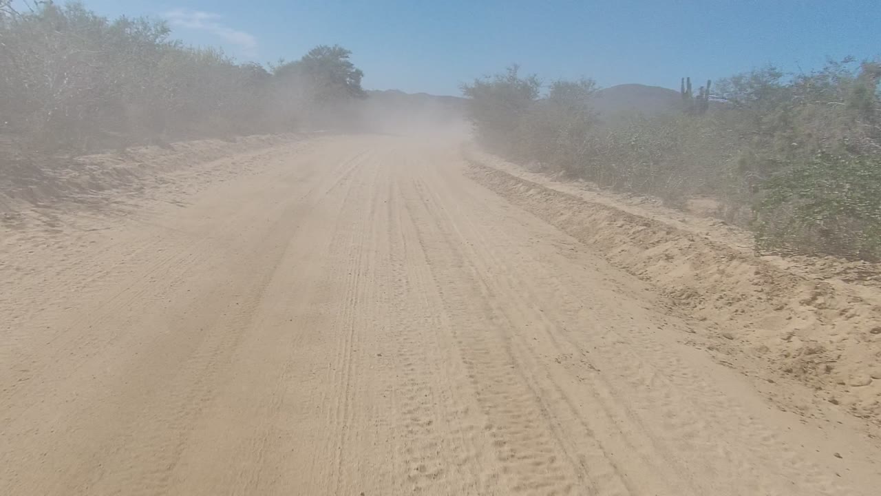 Centered view along a straight dirt road in Los Cabos, Baja California Sur, where roadside scrub and distant low hills are partly veiled by dust, with no visible people.