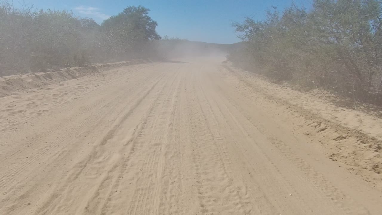 A sandy road in Los Cabos, Baja California Sur, bends gently through dry brush as a pale dust plume softens the background hills, with no visible people.