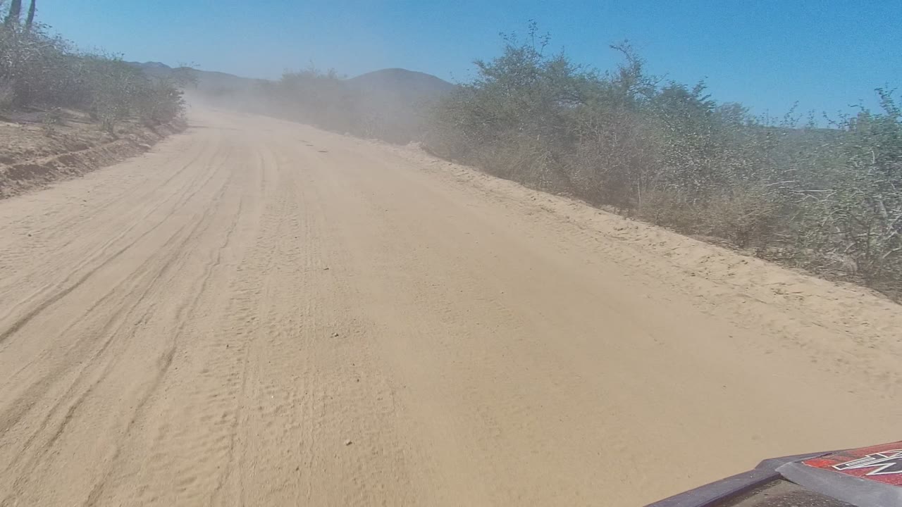 Forward vehicle-level view of a sandy dirt road in Los Cabos, Baja California Sur, with scrub bushes on both sides, faint hills ahead, a dust cloud in the distance, and no visible people.