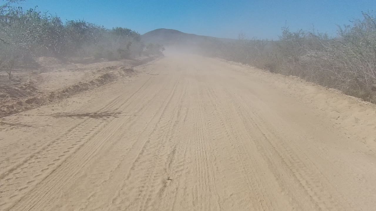 A sandy dirt road in Los Cabos, Baja California Sur stretches ahead between sparse desert shrubs, with dust over the track and no visible people.