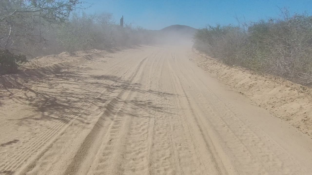 A Los Cabos sandy dirt road runs between sparse desert brush, with tire tracks in the foreground, a dust cloud farther ahead, distant hills, and no visible people.