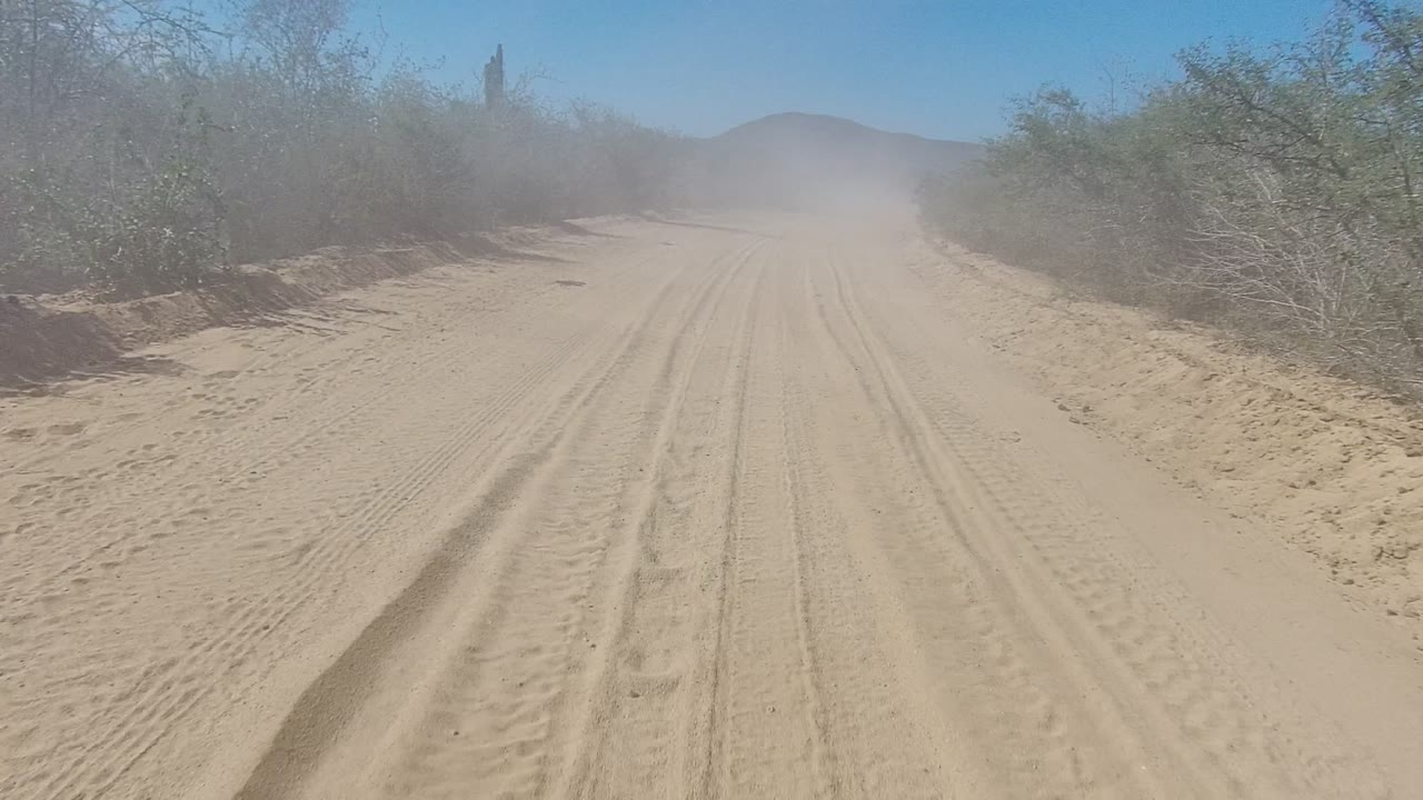 The Los Cabos road remains centered with parallel tire ruts, low scrub tight to both sides, haze from dust in the distance, and no visible people.