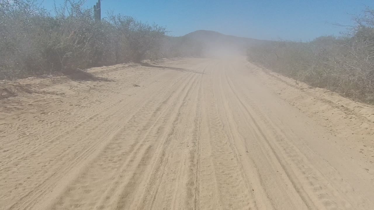 A wide Los Cabos desert track fills the frame, with sandy tire marks leading toward a dust cloud and low hills beyond, with no visible people.