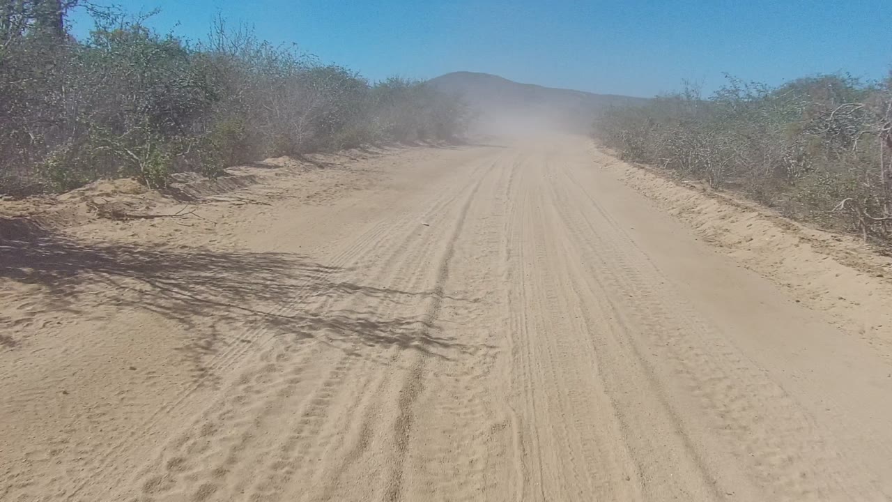 Sandy road curving slightly through desert brush with dust in the distance.