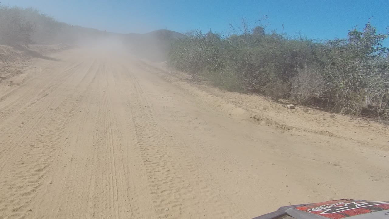 Start of the Los Cabos desert road clip, showing tire tracks in sand, roadside scrub, distant low hills, a dust cloud ahead, and no visible people.