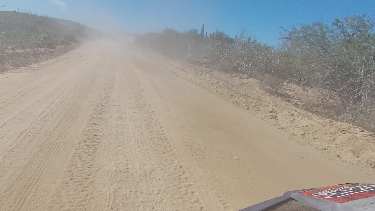 Opening frame of a sandy dirt road in Los Cabos, Baja California Sur, with tire tracks, desert shrubs, a blue sky, a dust cloud ahead, and no visible people.