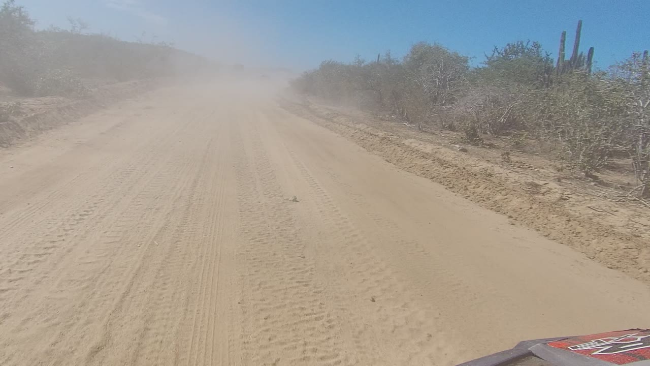 The vehicle view continues along a pale sandy road in Los Cabos, with visible track marks, cactus and scrub to the right, dust suspended in the distance, and no visible people.