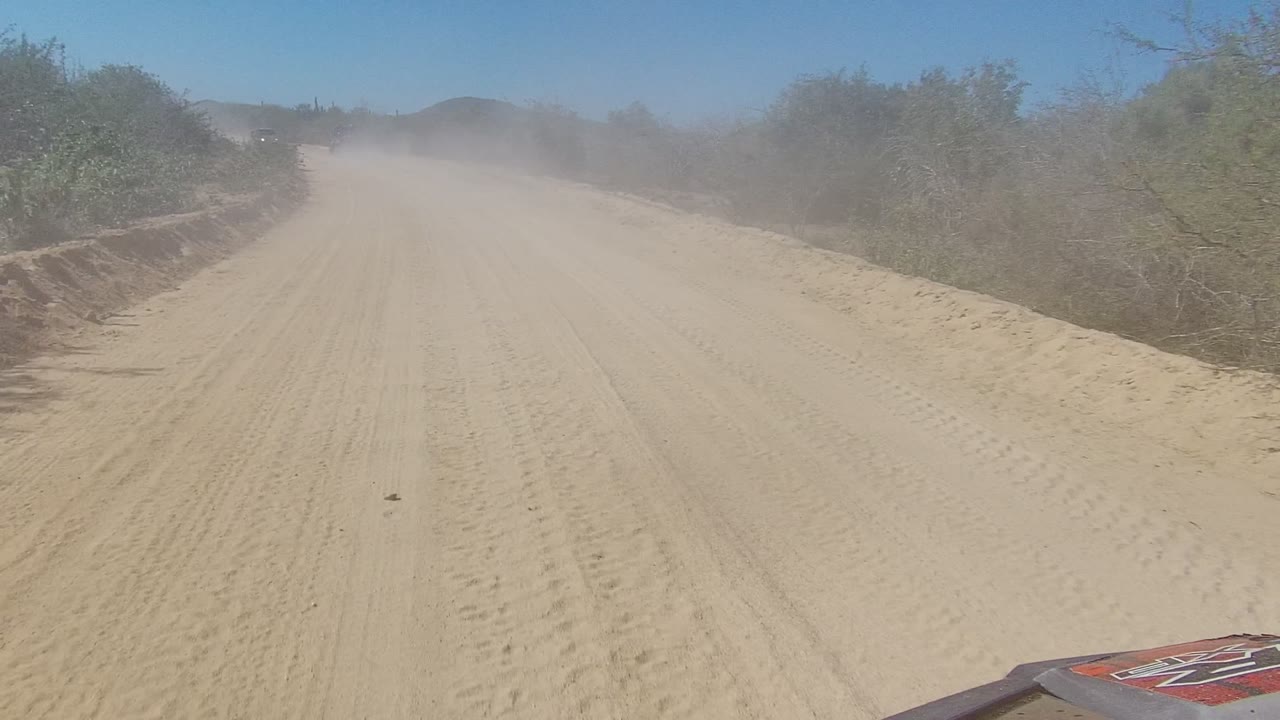 Sandy dirt road in Los Cabos, Baja California Sur with dry scrub on both sides, dust hanging ahead, low hills in the distance, and no visible people.