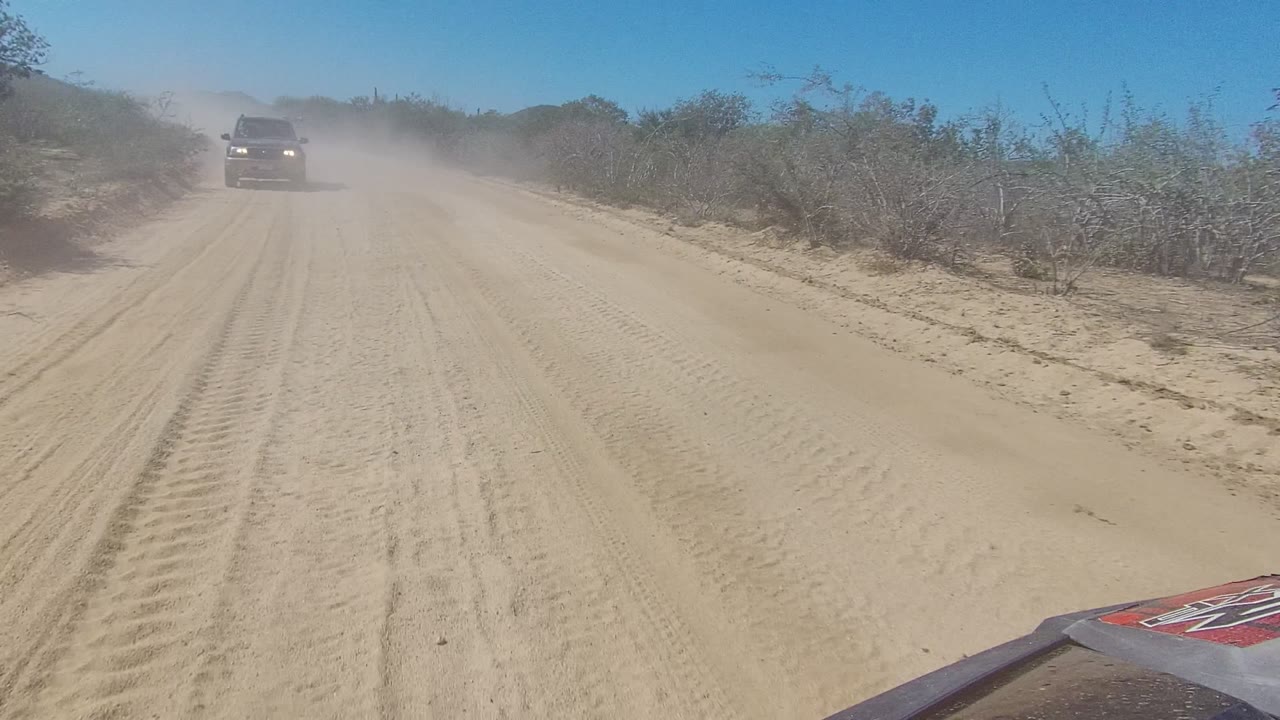 The lead vehicle sits near the center of a dusty Los Cabos dirt road, with tire tracks in the sand, brush on both sides, and no visible people.