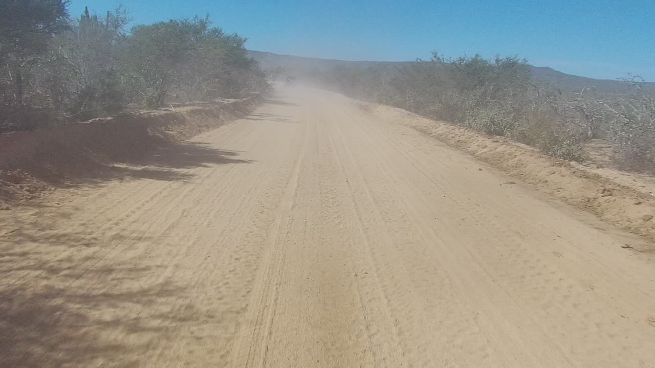 Opening frame of a sandy dirt road in Los Cabos, Baja California Sur, with dry brush on both sides, a faint dust cloud ahead, and no visible people.