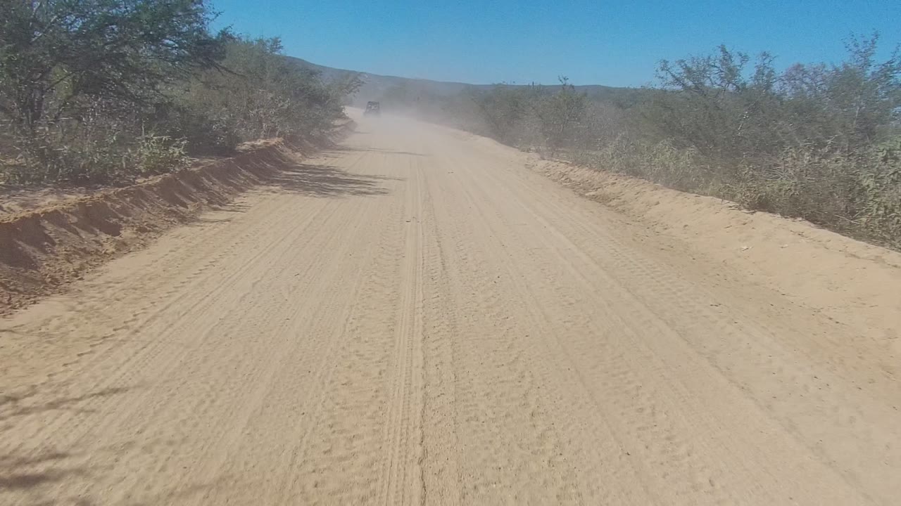 A forward road view in Los Cabos shows the sandy track narrowing toward a dust plume, with low scrub, tree shadows, distant hills, and no visible people.