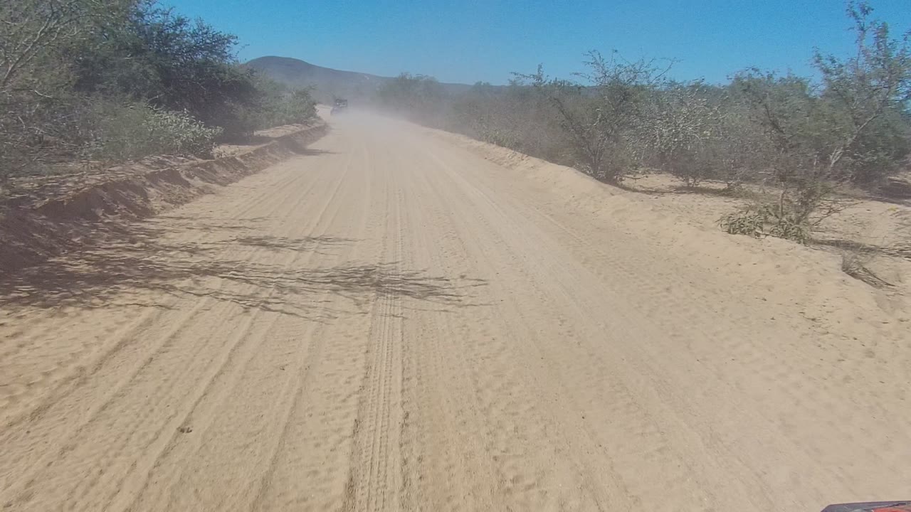 A distant vehicle appears near the center of the Los Cabos dirt road, partially obscured by airborne dust, with dry shrubs and low hills behind it and no visible people.