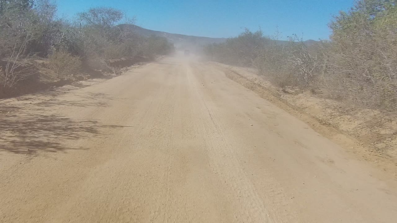 A wider view of the Los Cabos dirt road shows shrub-lined shoulders, pale sand, dust drifting in the distance, and no visible people.