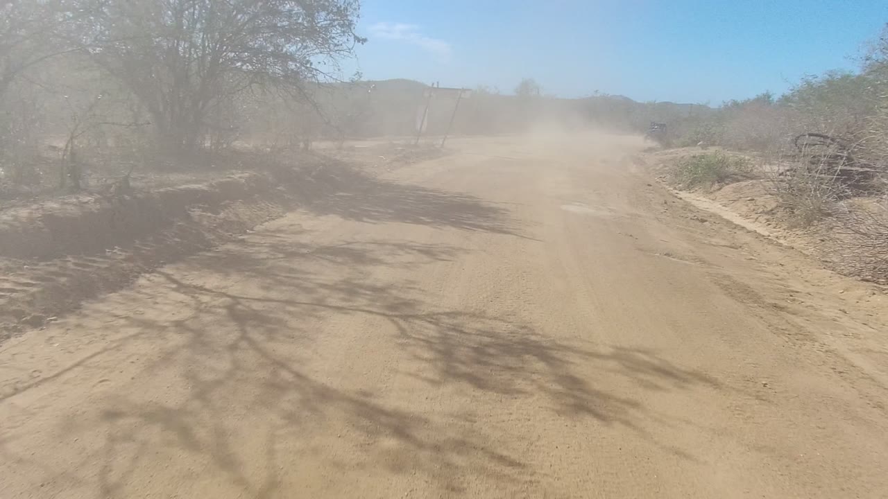 A broad dirt road in Los Cabos, Baja California Sur runs past leafless brush and cactus while a dust cloud drifts across the track, with no visible people.