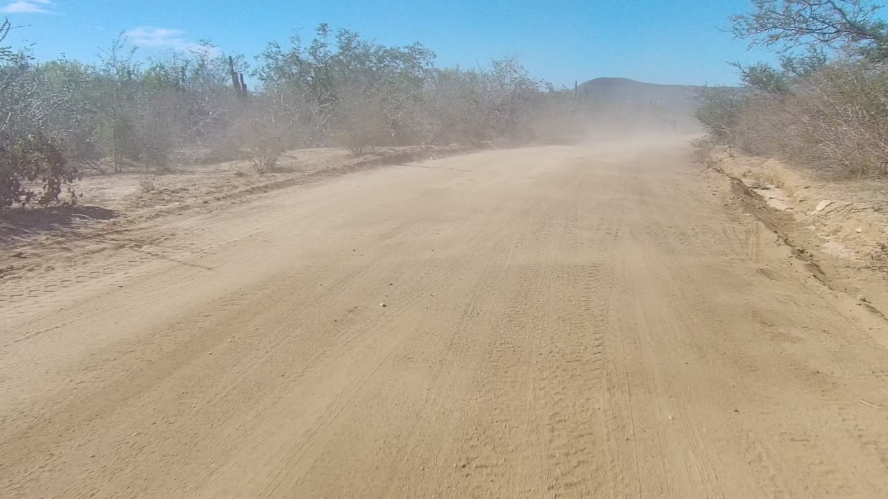 A sandy dirt road in Los Cabos, Baja California Sur runs between sparse shrubs, with a dust cloud hanging ahead and no visible people.