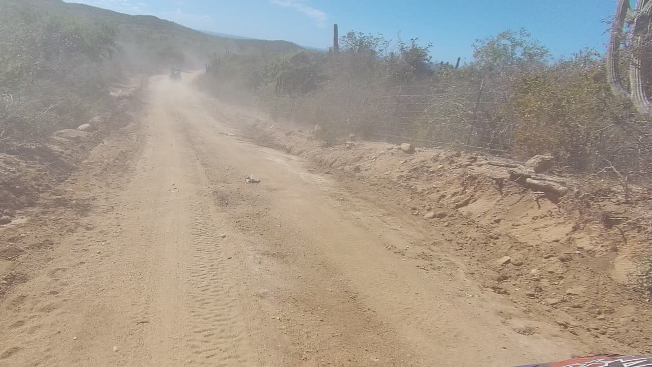 A dusty rural dirt road in Los Cabos, Baja California Sur stretches toward low hills with dry scrub on both sides and no visible people.