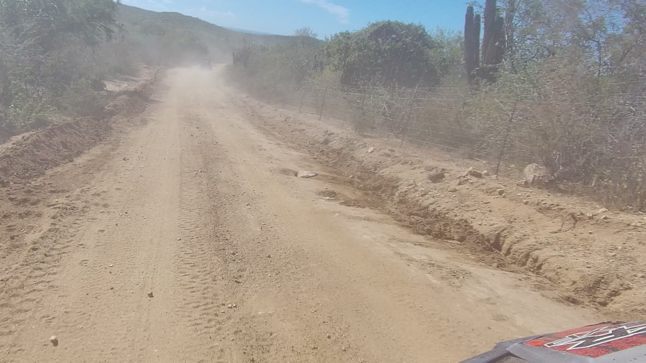 Straight dirt road with dust plume, scrub, cactus, and fence line.