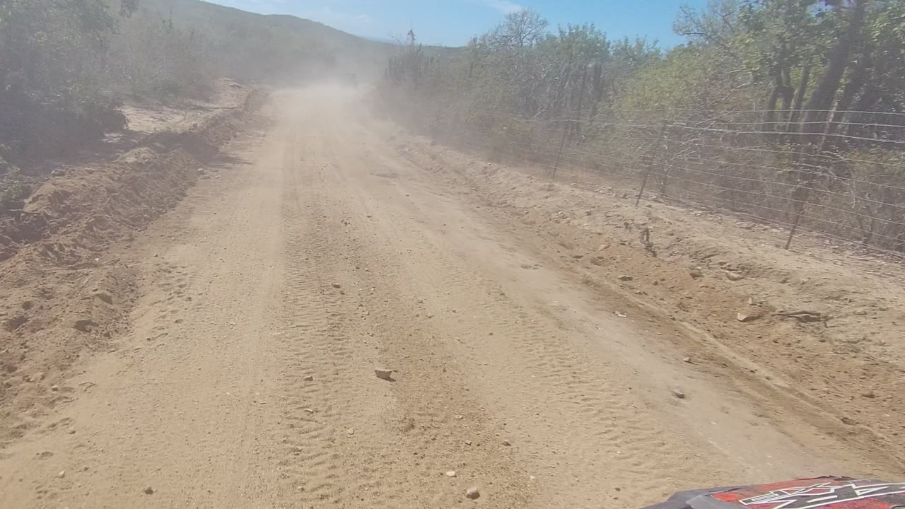A pale dirt road in Los Cabos shows tire tracks and loose rocks, with a roadside ditch, wire fencing, dry vegetation, and no visible people.