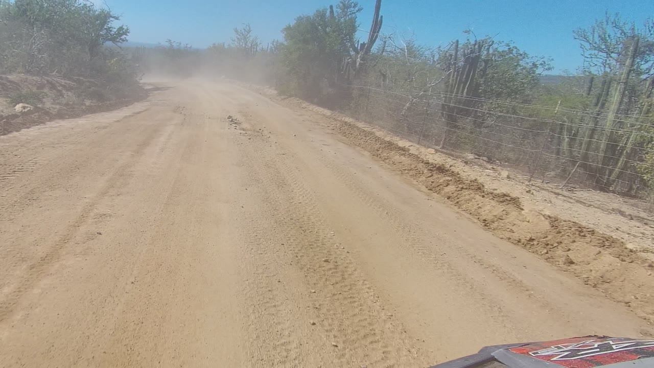 Opening frame of a dusty dirt road in Los Cabos, Baja California Sur, with cacti, sparse bushes, a wire fence on the right, and no visible people.