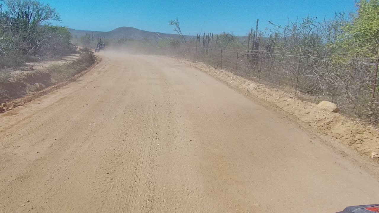 Low road view in Los Cabos showing a dusty dirt track, desert bushes and cacti, a distant vehicle ahead, and no visible people.