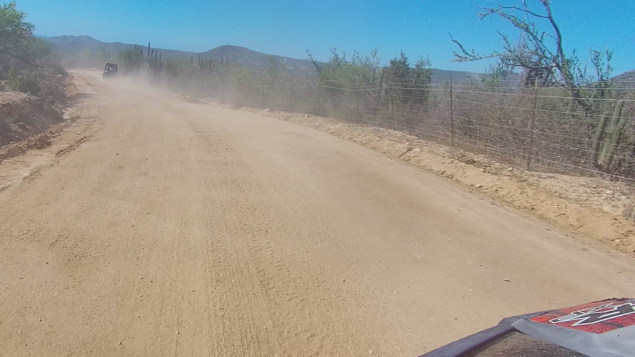 A later moment on the Los Cabos road shows the vehicle farther ahead in dust, sandy shoulders, thorny vegetation, cacti, distant hills, and no visible people.