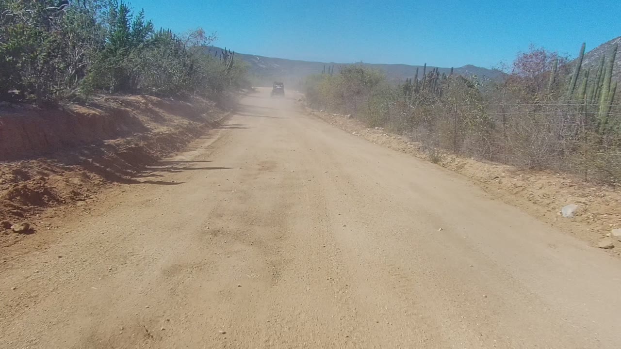 A dusty dirt road in Los Cabos, Baja California Sur stretches toward a small distant vehicle between dry shrubs, cactus, wire fencing, and low hills, with no visible people.