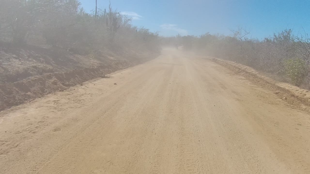 A wider view of the sandy road in Los Cabos shows dust suspended over the far lane, desert scrub and cactus silhouettes along both sides, and no visible people.