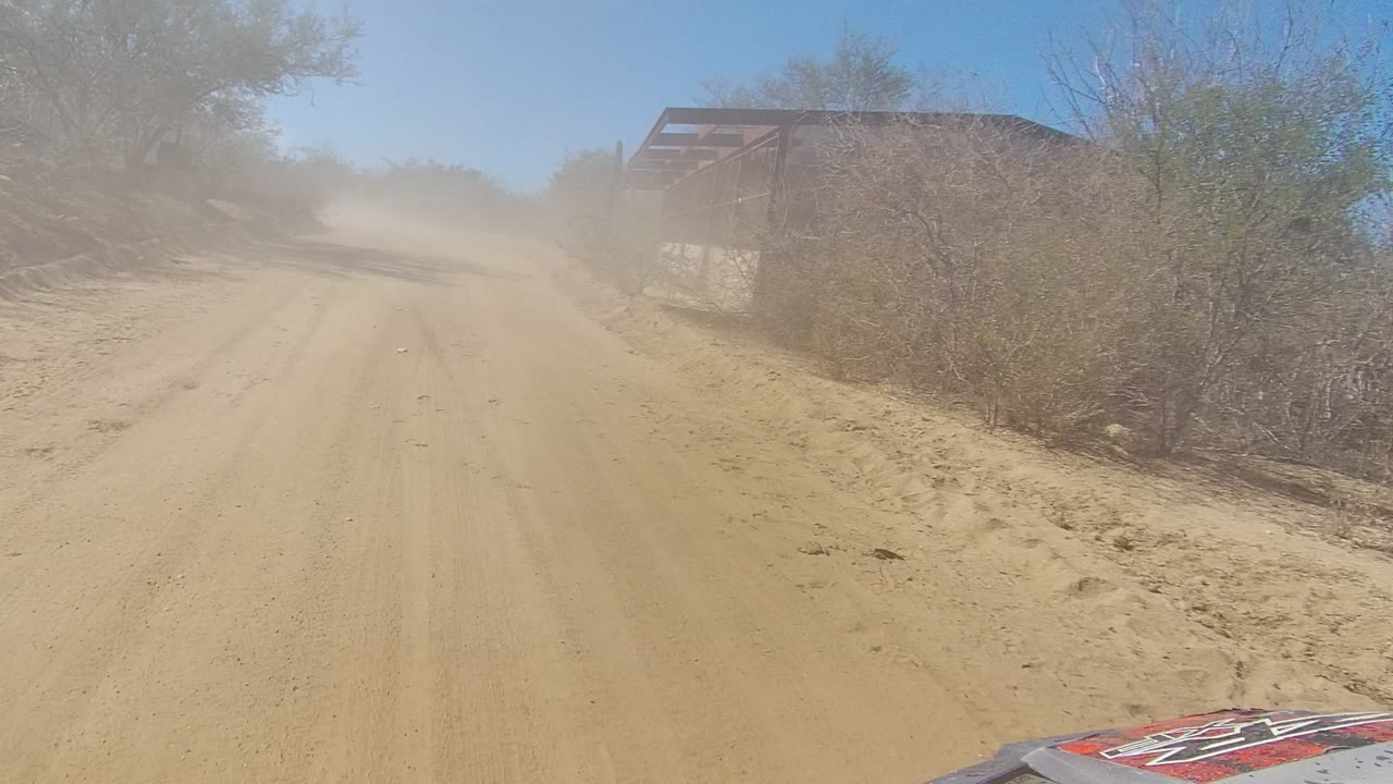 At 0:00 in Los Cabos, a sandy dirt road runs past dry shrubs under a clear blue sky, with a dust cloud ahead and no visible people.