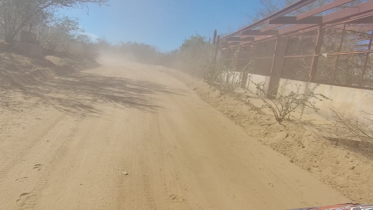 At 0:01 in Los Cabos, the vehicle passes a red metal-framed roadside structure with cactus and scrub along the sandy road, and no visible people.