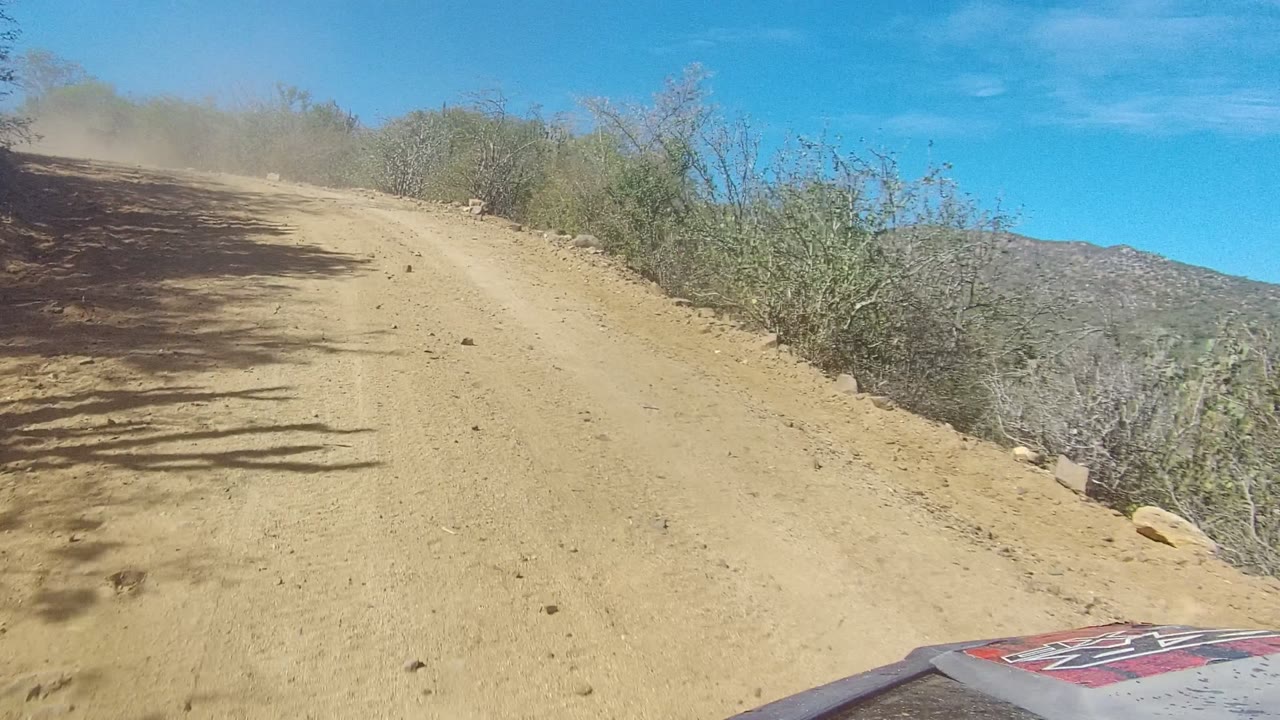 Start of the Los Cabos dirt road view, with tan road surface, sparse roadside brush, blue sky, vehicle hood at the lower right, and no visible people.