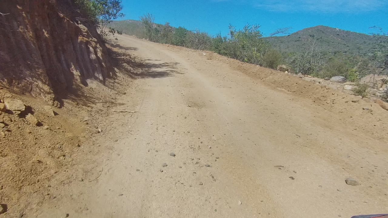 Vehicle POV at the start of a sandy uphill dirt road in Los Cabos, Baja California Sur, with dry vegetation on both sides, distant hills, blue sky, and no visible people.