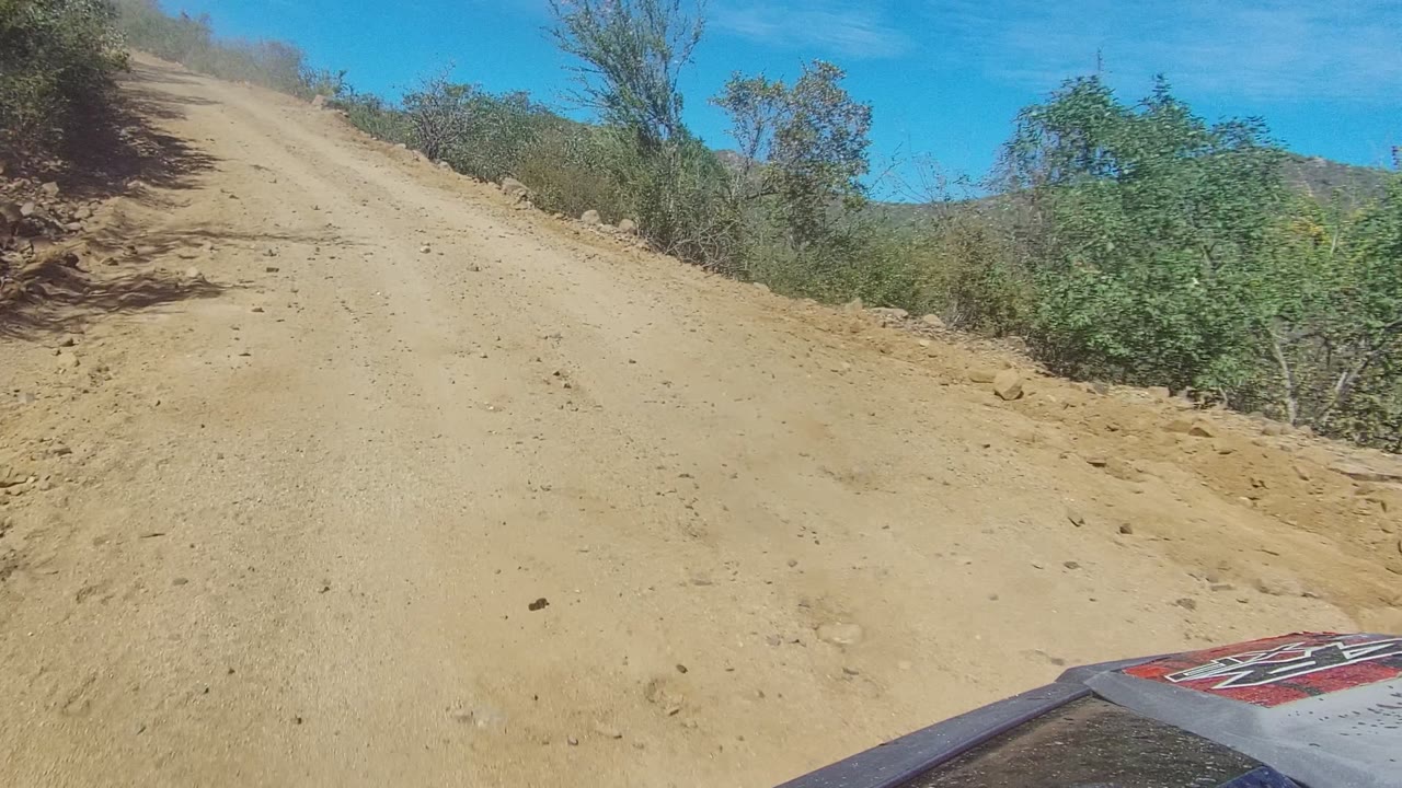 The vehicle advances toward a sandy rise in Los Cabos, with brush lining the road, hills beyond the right edge, a visible vehicle corner, and no visible people.