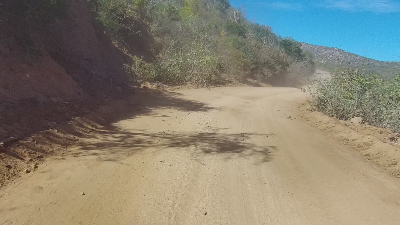 Sandy dirt road in Los Cabos curves along a brushy hillside with dust ahead and no visible people.