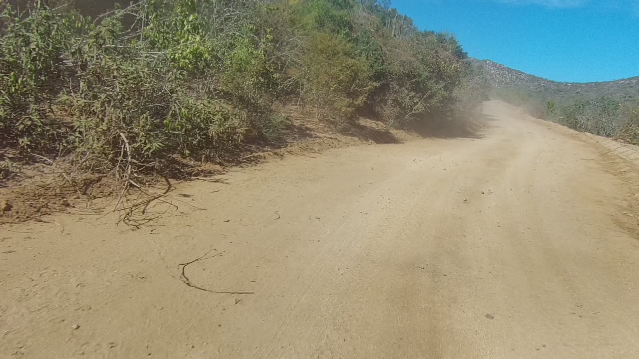 Los Cabos dirt road runs forward between dense dry vegetation and a distant hill, with a light dust plume and no visible people.