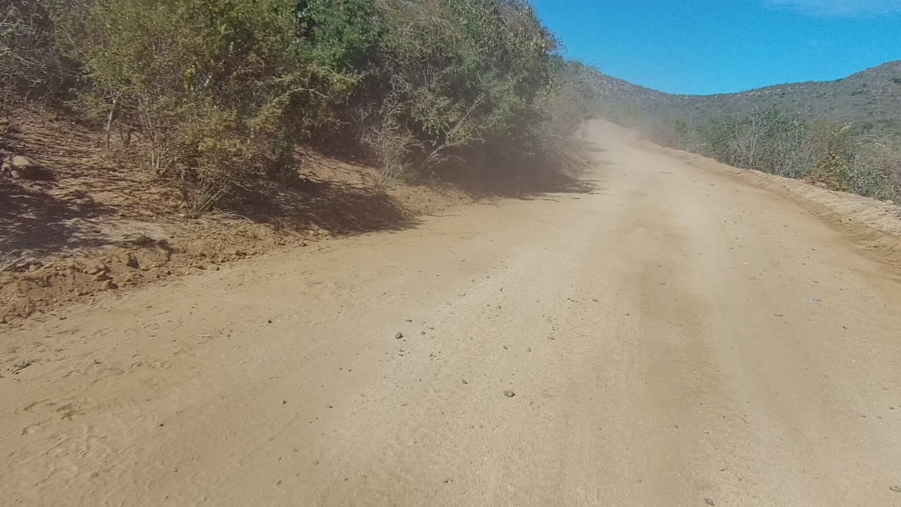 A sandy road in Los Cabos rises gently through scrub-covered terrain with dust in the distance and no visible people.