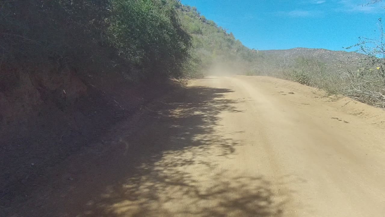 Dust plume over a dirt road curving through brushy hills.