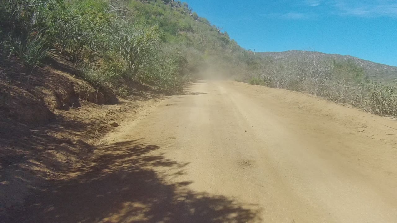 A wider section of the Los Cabos dirt road bends gently right, bordered by green-gray scrub, exposed dry soil, distant rocky hills, bright blue sky, and no visible people.