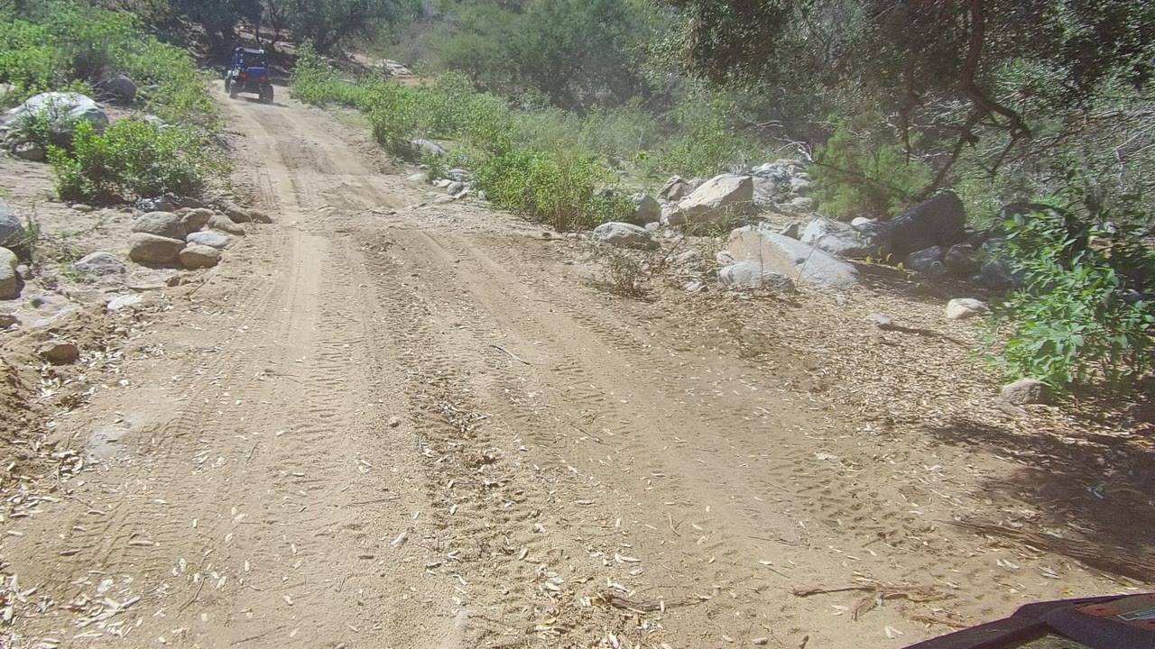 A dusty Los Cabos dirt road fills the frame, bordered by pale rocks and green scrub, with the off-road vehicle small in the distance and no visible people.
