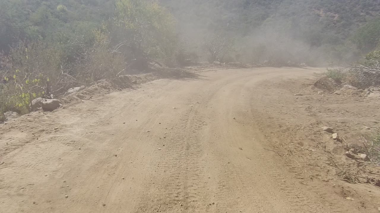 The road in Los Cabos appears to split or bend around dry brush, with dusty air, rocky shoulders, and a small vehicle hood edge visible at the bottom, with no visible people.