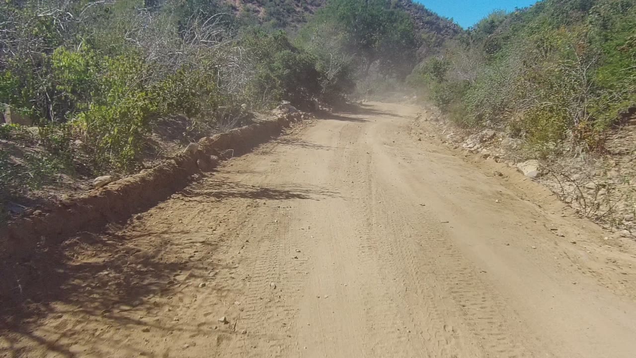 Forward view down a dusty Los Cabos dirt road bordered by dry scrub, rocks, and low hills, with no visible people.