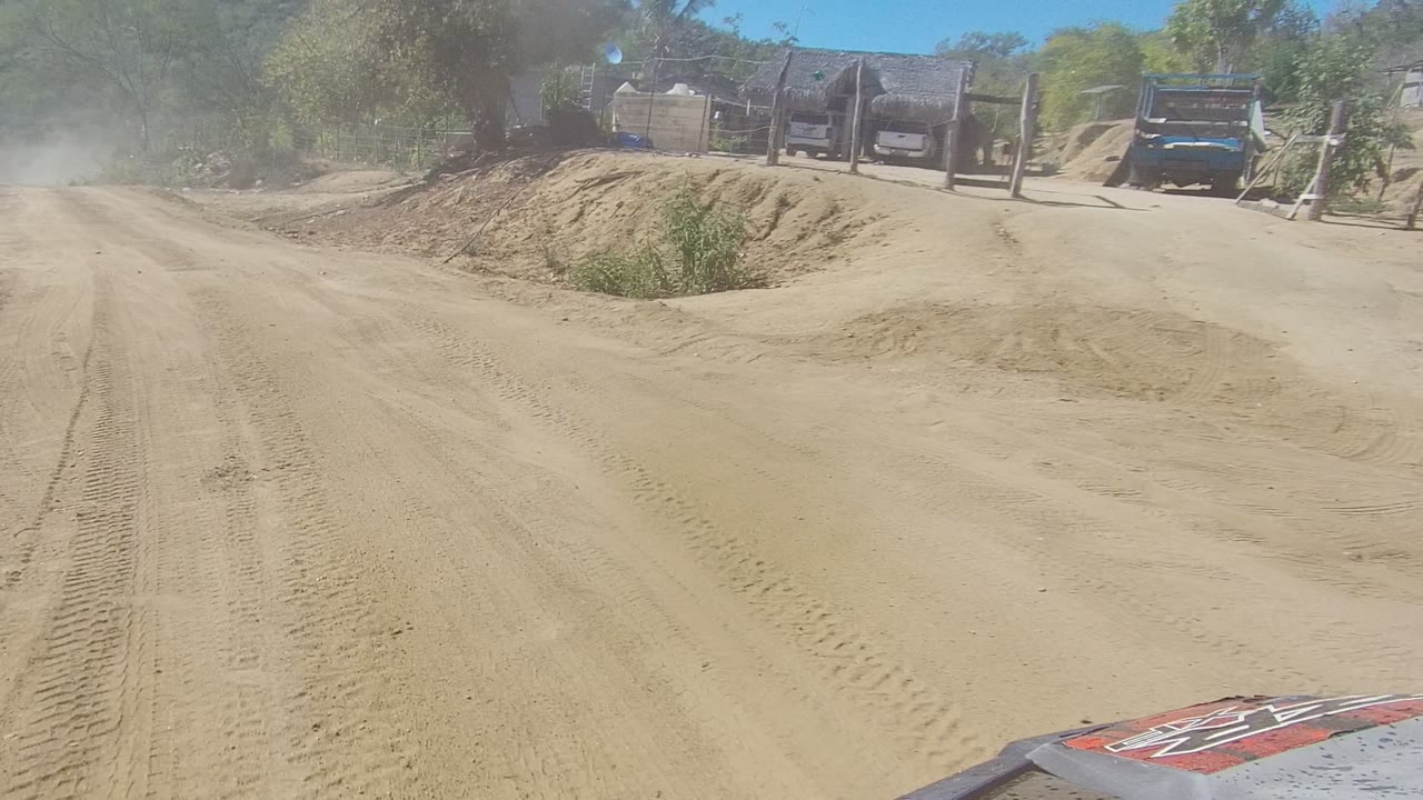 A straight stretch of dirt road in Los Cabos shows tire tracks, roadside fencing, dust in the air, trees, and the edge of the vehicle hood, with no visible people.