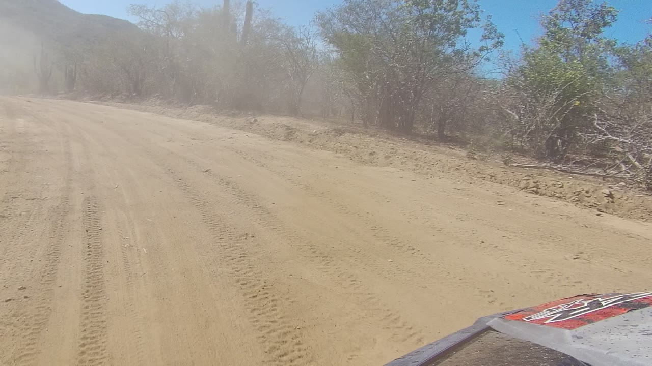 The road runs straighter through Los Cabos desert scrub, with tire lines in the sand, dust suspended ahead, cactus silhouettes on both sides, distant hills, and no visible people.